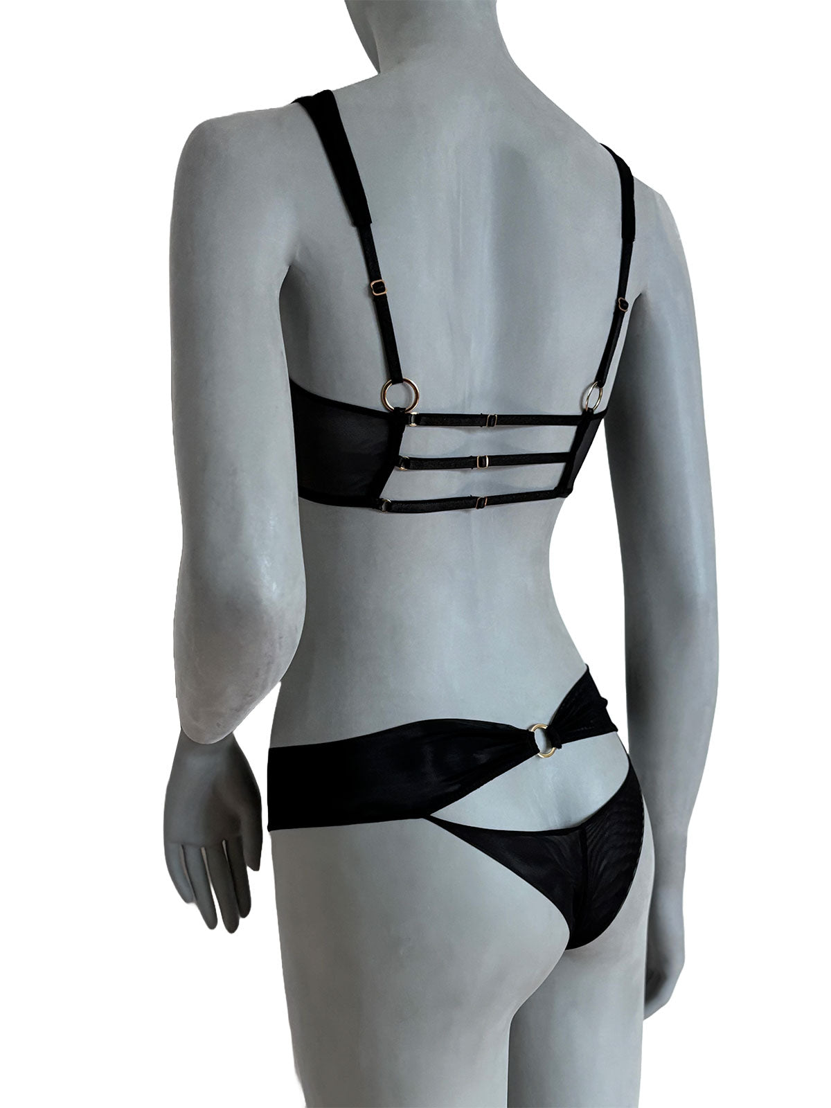 Sutiã Julie preto em tule transparente com alças largas pretas ajustáveis, três tiras horizontais em tule com detalhes em argola banhada a ouro, calcinha preta em tule com tiras cruzadas, vista costas em manequim fundo neutro, conjunto lingerie sem bojo design autoral Cisô Atelier