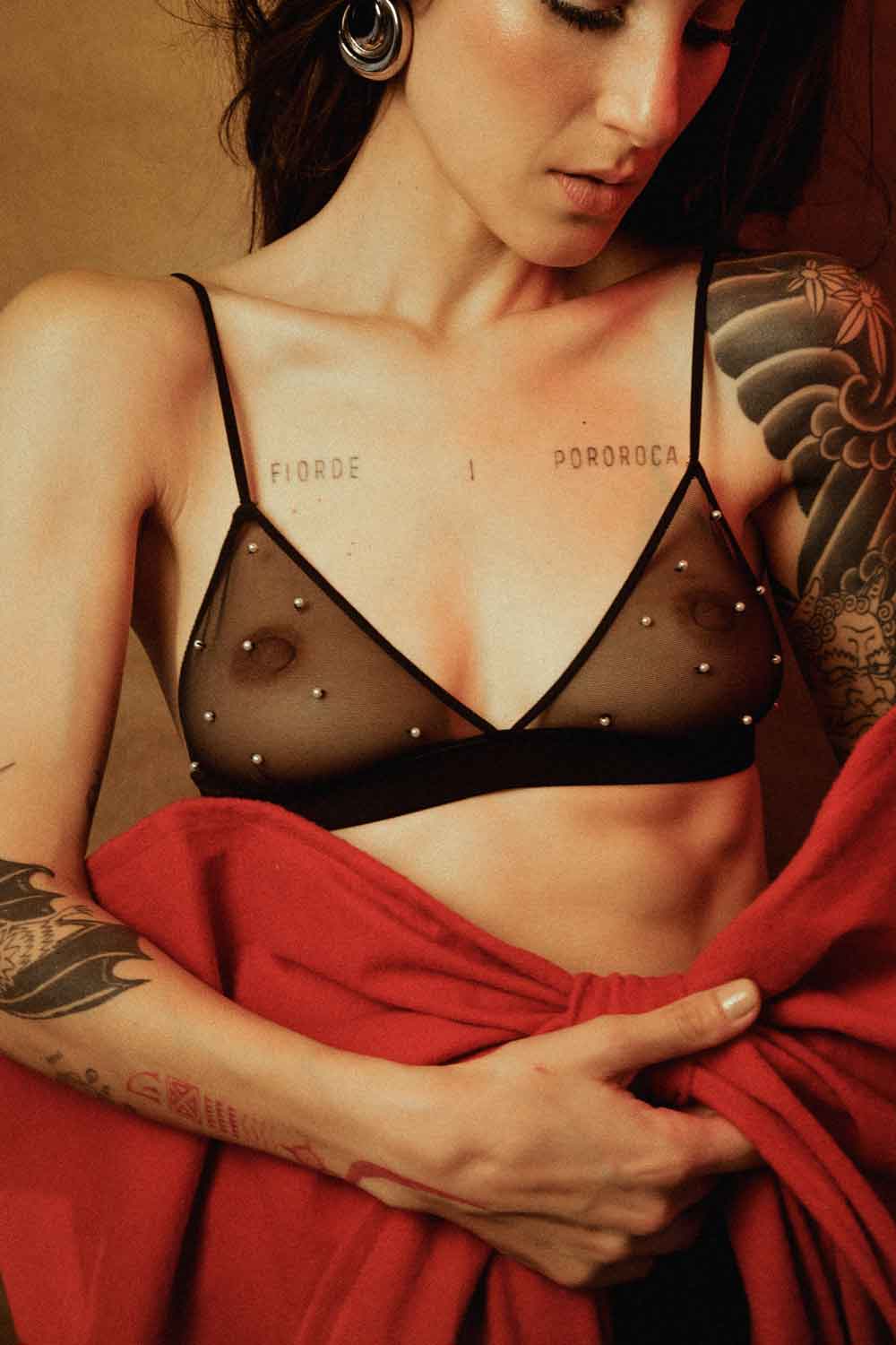 Mulher usando sutiã de tule preto com pérolas Cisô Atelier, segurando tecido vermelho em forma de laço, em clima natalino sofisticado e sensual